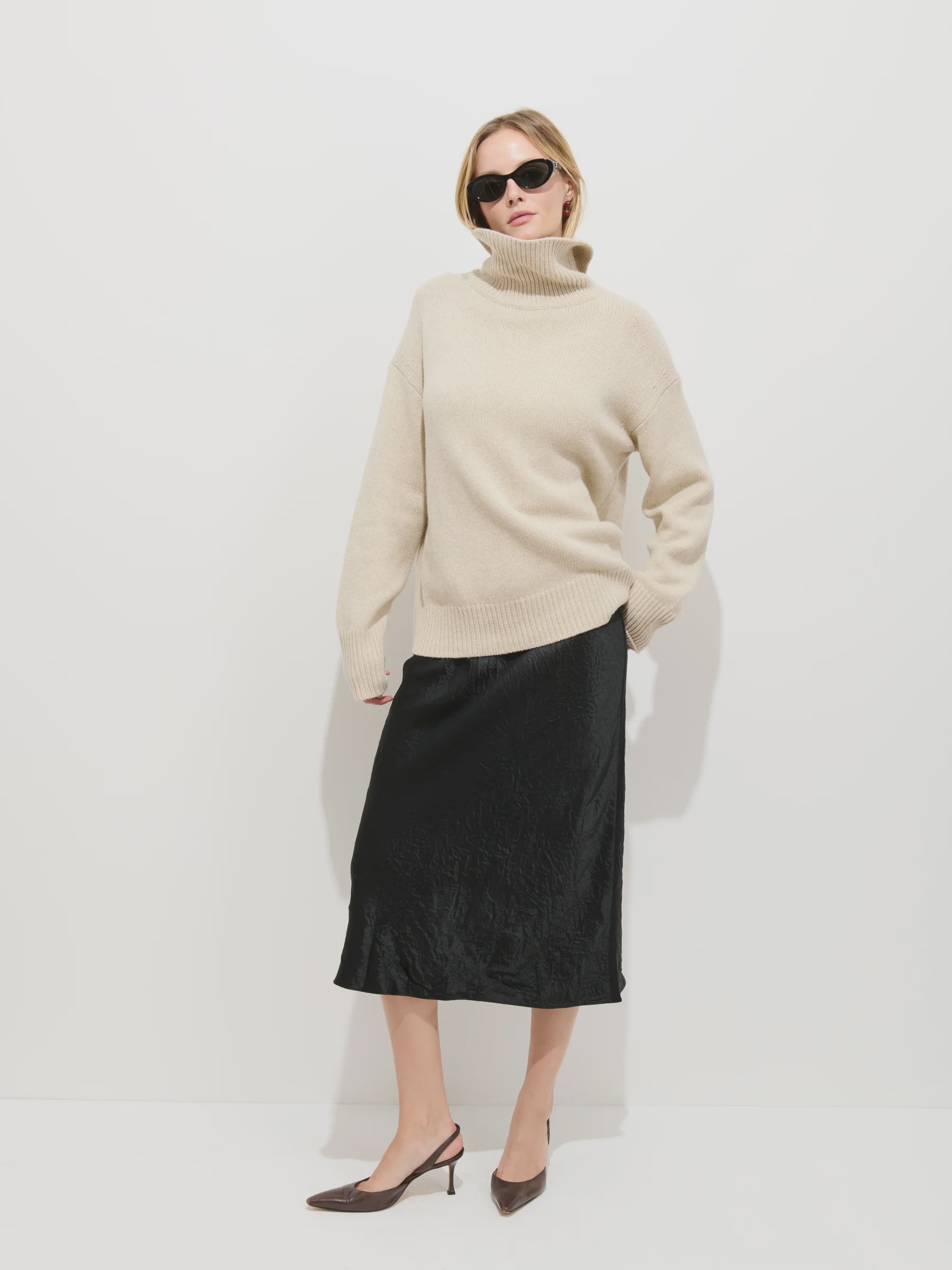 A woman stands against a white background wearing sunglasses, a beige turtleneck, brown heeled shoes, and the Alex Mill Claire Skirt—a black bias cut pull-on skirt.
