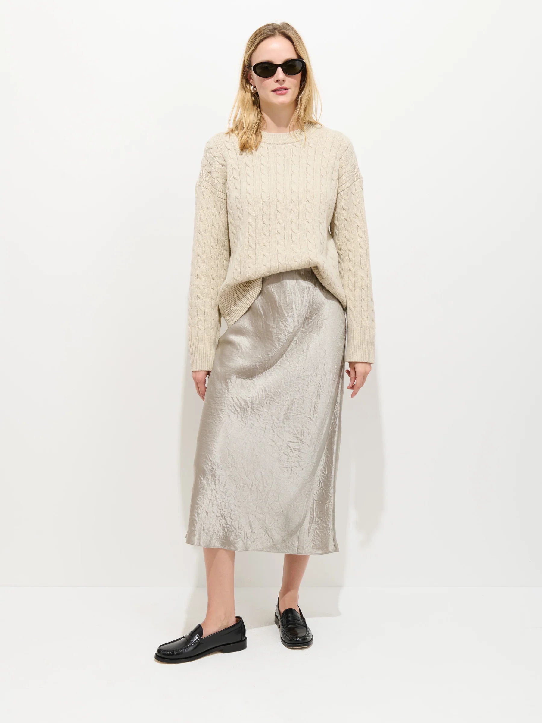 A woman with blonde hair and a neutral expression stands against a white background wearing the Claire Skirt by Alex Mill, a cream cable-knit sweater, black loafers, and dark sunglasses.