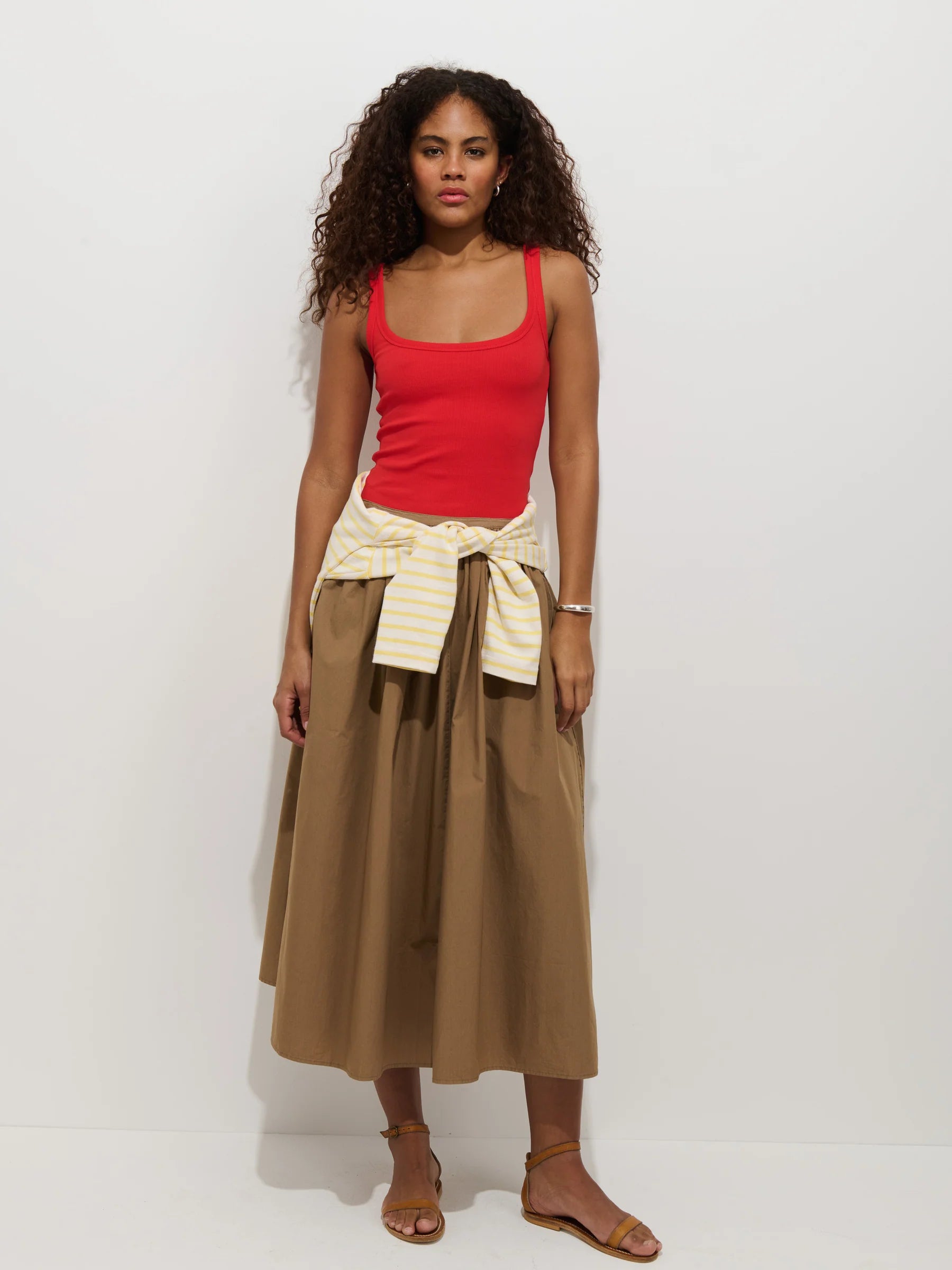 A woman with curly hair wears a red tank, tan sandals, a yellow-and-white striped sweater tied at her waist, and the Alex Mill Sarah Skirt—a brown cotton poplin midi skirt with an invisible zipper—while standing against a plain white background.