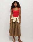 A woman with curly hair wears a red tank, tan sandals, a yellow-and-white striped sweater tied at her waist, and the Alex Mill Sarah Skirt—a brown cotton poplin midi skirt with an invisible zipper—while standing against a plain white background.