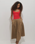 A woman with curly hair wears the Alex Mill Sarah Skirt, a high-waisted, tan, ankle-length cotton skirt with pockets. She stands against a white background, looking down with her hands in her skirt pockets.