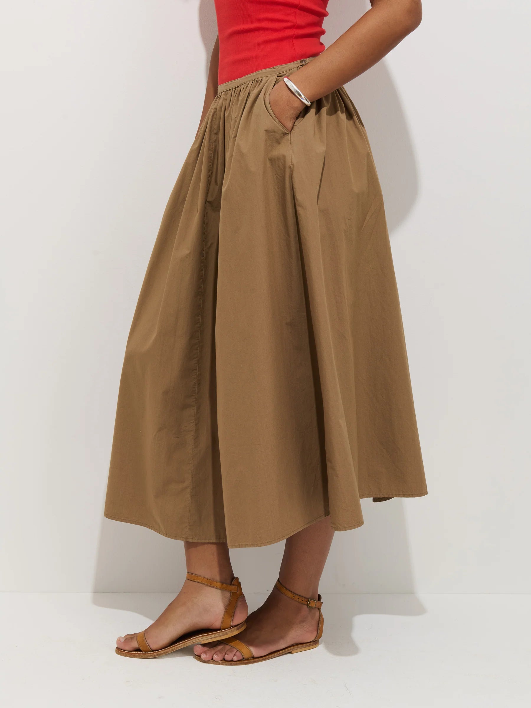 A person wearing the Alex Mill Sarah Skirt—a tan, high-waisted cotton poplin midi skirt with pockets and an invisible zipper—paired with a red sleeveless top and brown flat sandals, stands against a plain white background.