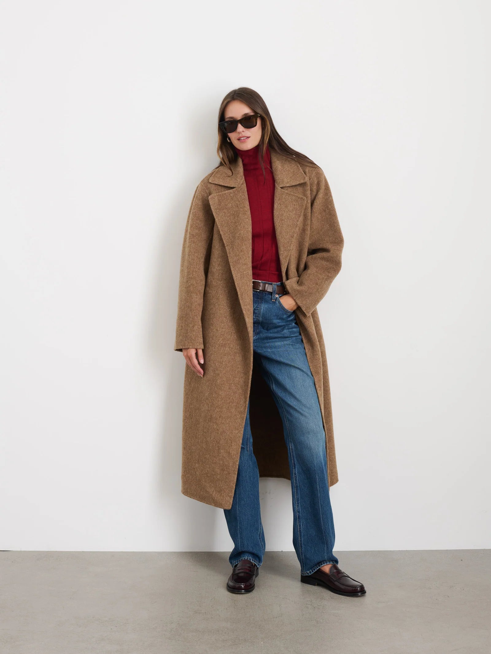 A person wearing sunglasses stands against a white wall, dressed in an Alex Mill Georgia Wrap Coat, a red turtleneck, blue jeans, and brown loafers. They have long hair and are casually posing with one hand in their pocket.