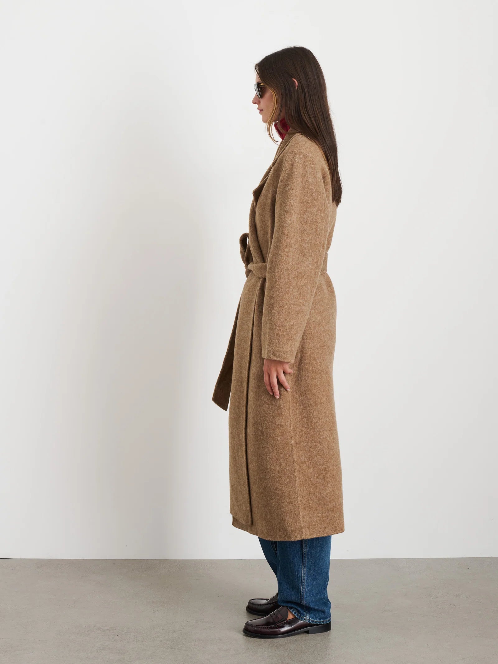 A person in a Georgia Wrap Coat by Alex Mill and blue jeans stands sideways against a plain white background. They are wearing sunglasses and dark shoes, with the coat belted at the waist.