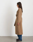 A person in a Georgia Wrap Coat by Alex Mill and blue jeans stands sideways against a plain white background. They are wearing sunglasses and dark shoes, with the coat belted at the waist.