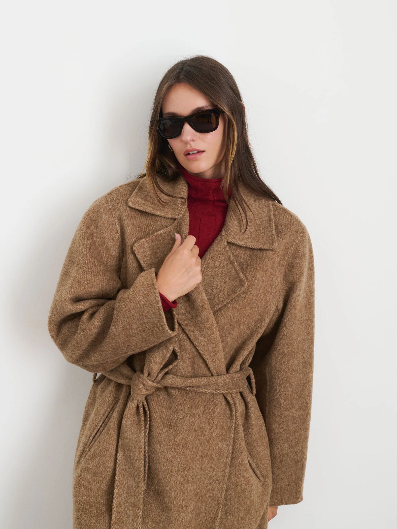 A person styled in the Alex Mill Georgia Wrap Coat, a long, brown vintage piece, poses against a plain white background. They wear it over a red top and complete their fashionable look with stylish sunglasses and long hair.