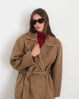 A person styled in the Alex Mill Georgia Wrap Coat, a long, brown vintage piece, poses against a plain white background. They wear it over a red top and complete their fashionable look with stylish sunglasses and long hair.