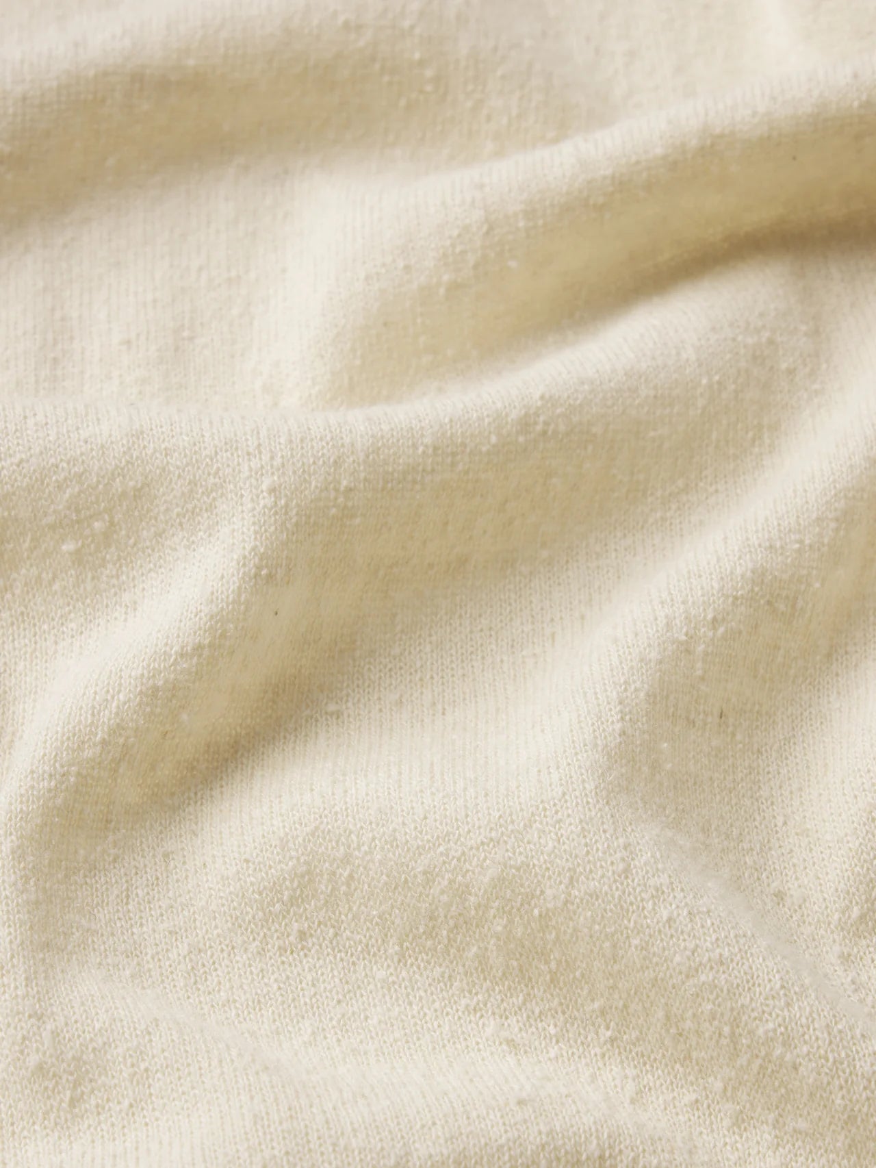 A close-up of The Knit Plus One Tee by FRAME shows its cream-colored, textured fabric with gentle folds and a nubby surface, capturing the relaxed luxury of this silk-inspired tee.