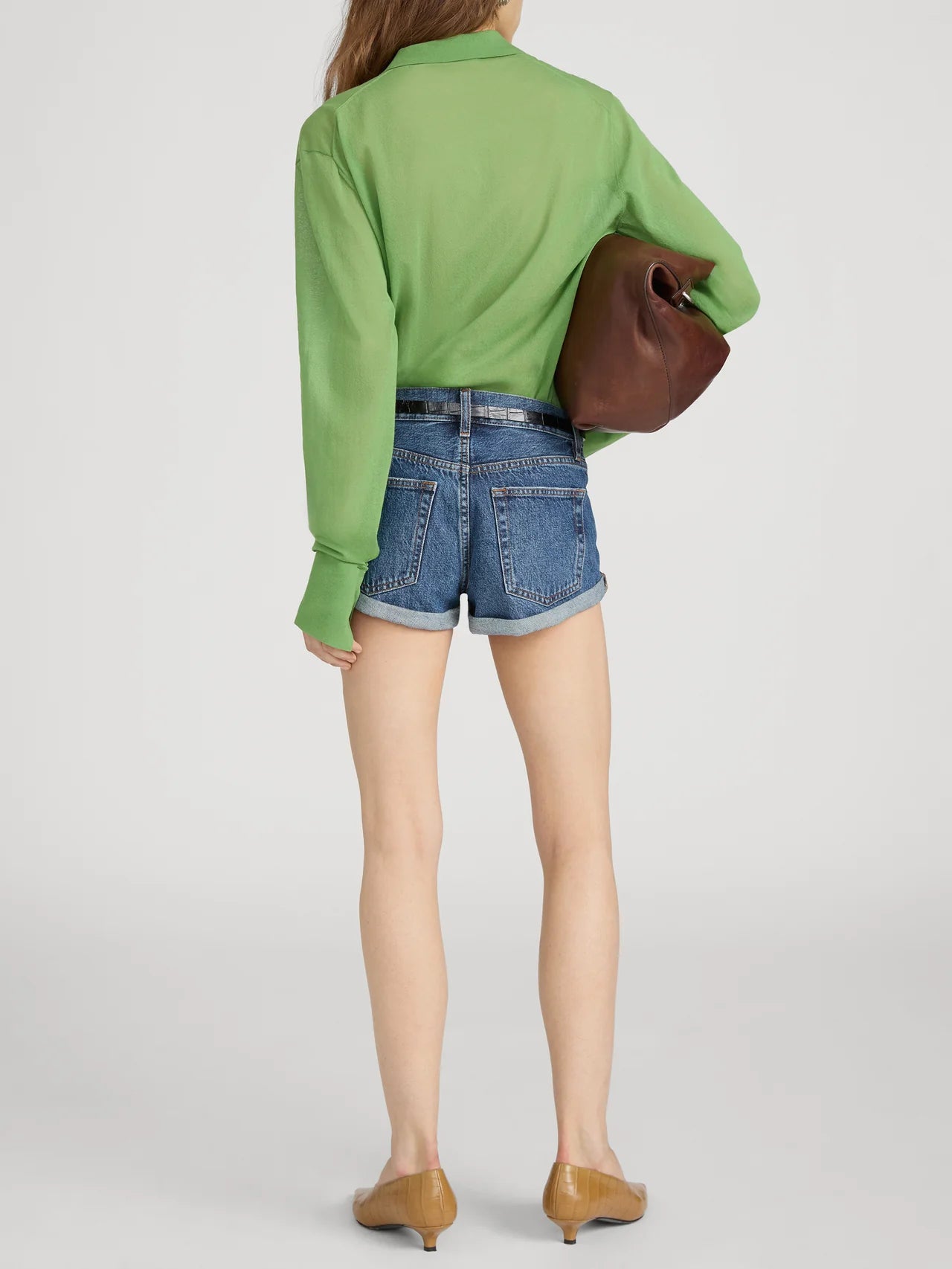 A person stands facing away, wearing FRAME’s The Collared Cardi in green with a slim fit, blue denim shorts, tan low-heeled shoes, and carrying a large brown clutch under one arm.