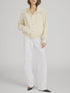 A person wearing FRAME's The Seersucker Track Jacket in light beige, paired with white wide-leg pants and black pointed-toe shoes, stands against a plain light gray background with their left hand in their pocket.