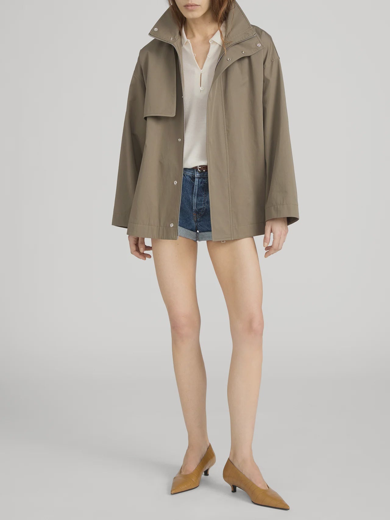 A woman models FRAME's The Short Field Trench in light brown over a white blouse, denim shorts, and brown low-heeled shoes against a plain background; her face is partially out of frame.