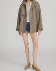 A woman models FRAME's The Short Field Trench in light brown over a white blouse, denim shorts, and brown low-heeled shoes against a plain background; her face is partially out of frame.