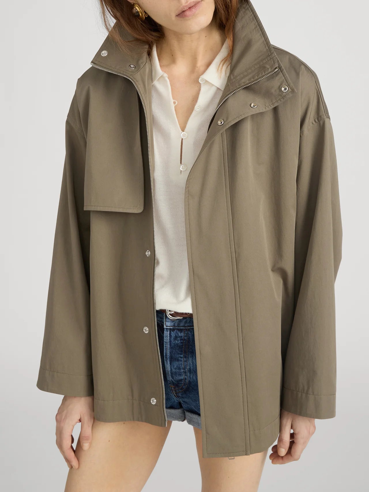 A woman layers FRAME's The Short Field Trench in olive green over a cream button-up shirt and denim shorts, standing against a plain background. Her face is partially cropped, highlighting effortless transitional style.