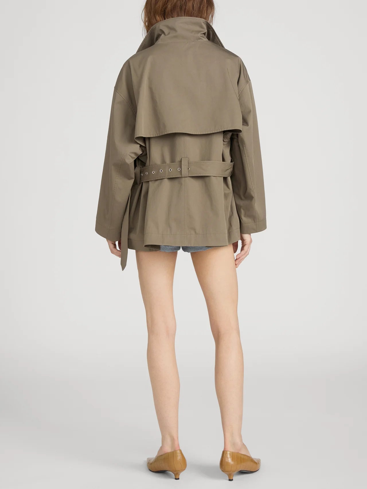 A person faces away, wearing FRAME's The Short Field Trench with denim shorts and tan flats—demonstrating effortless transitional layering against a plain white background.