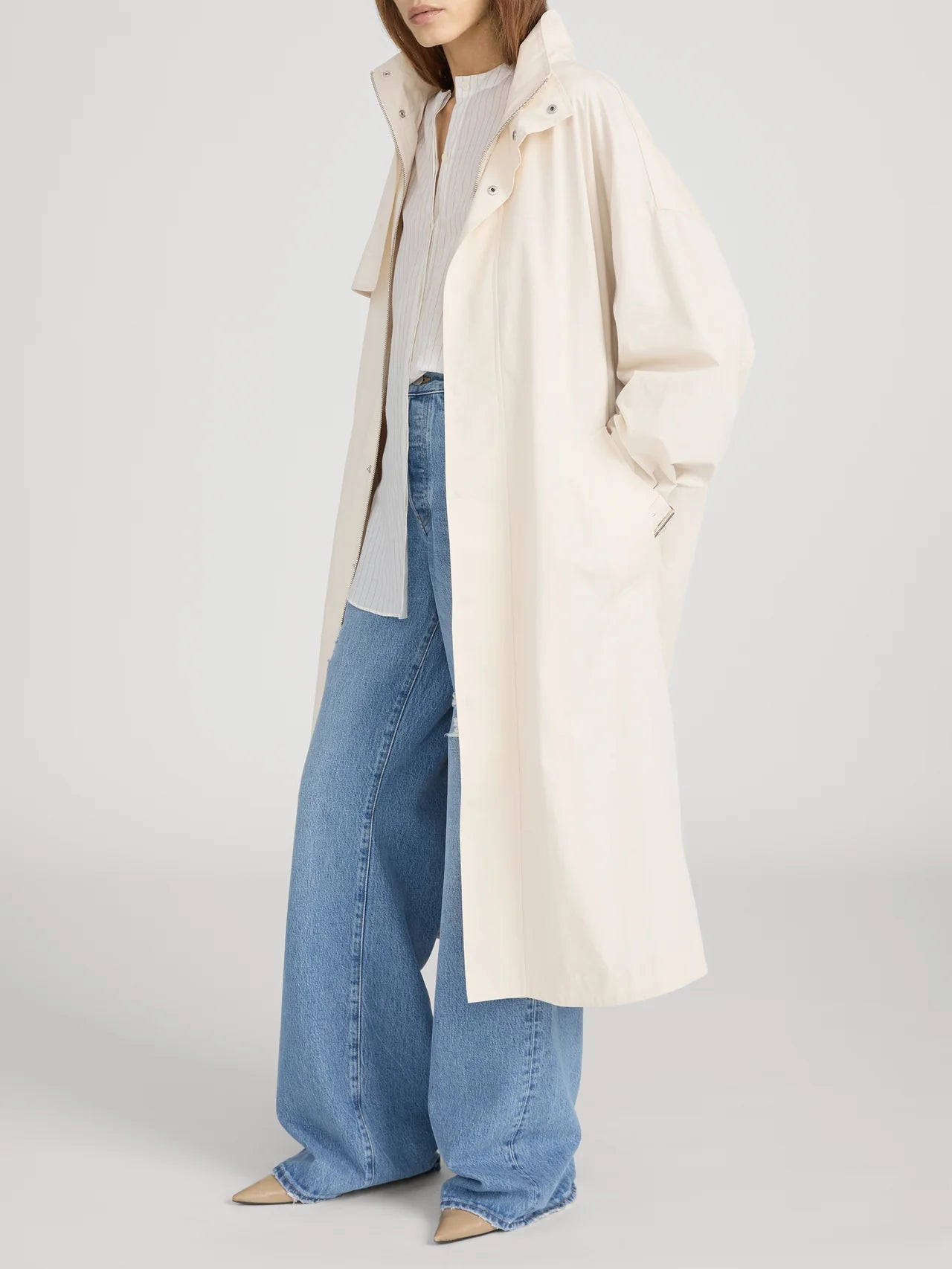 A woman wears The Field Trench by FRAME—a long, loose, cream cotton coat—over a light shirt and wide-leg blue jeans with beige pointed-toe shoes. One hand is in her pocket and her head is turned slightly to the side.