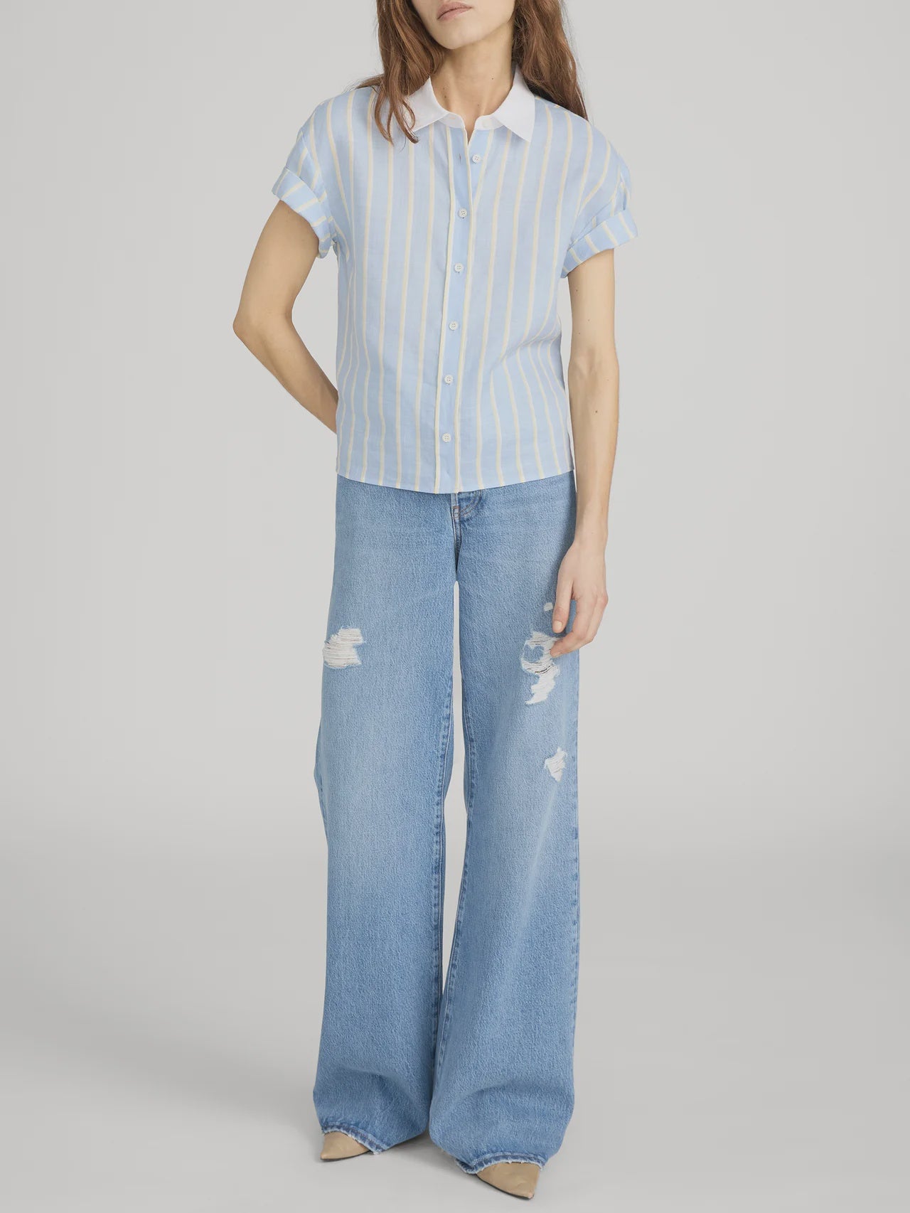 A person wears FRAME's The Boardwalk Shirt, a short-sleeve light blue and white striped linen top, paired with wide-leg light blue ripped jeans and beige shoes, standing against a plain gray background.