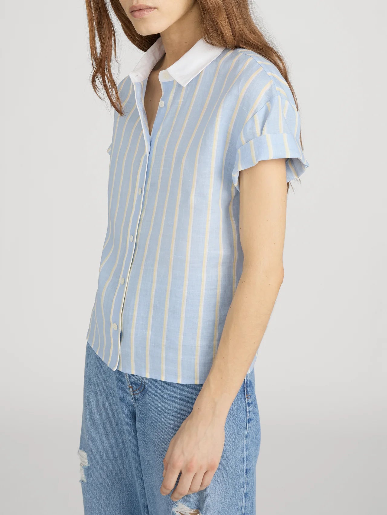 A woman wearing FRAME's The Boardwalk Shirt—a relaxed fit, light blue shirt with white and yellow stripes and a white collar—paired with light blue ripped jeans stands against a plain background.