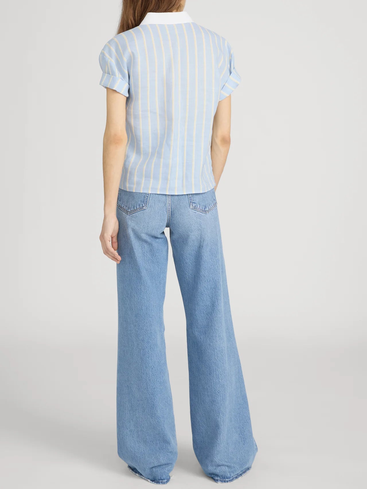 A person stands with their back to the camera, wearing FRAME’s The Boardwalk Shirt in a relaxed fit with white and yellow stripes, paired with high-waisted, wide-leg blue jeans. The background is plain white.