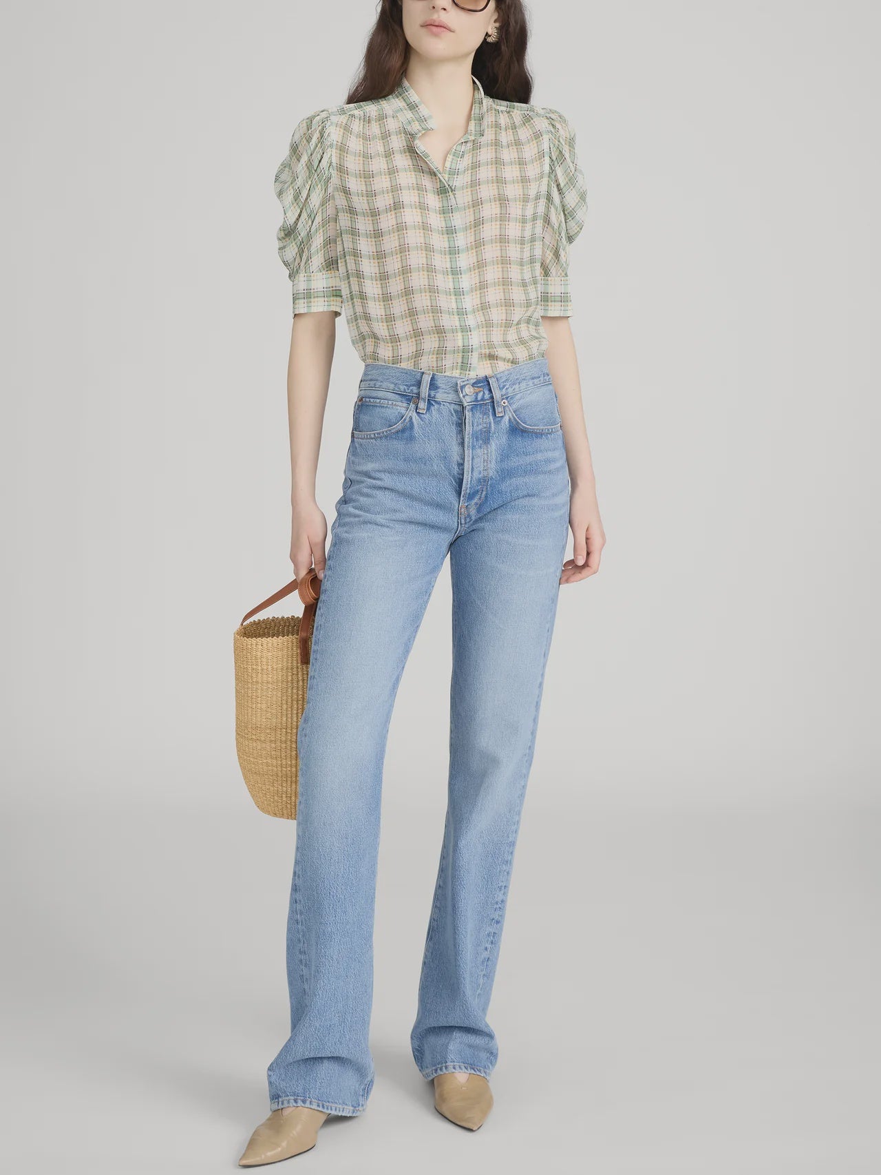 A woman stands in a neutral setting wearing FRAME's The Gillian Top, styled with high-waisted light blue jeans, beige pointed shoes, and carrying a woven basket bag.