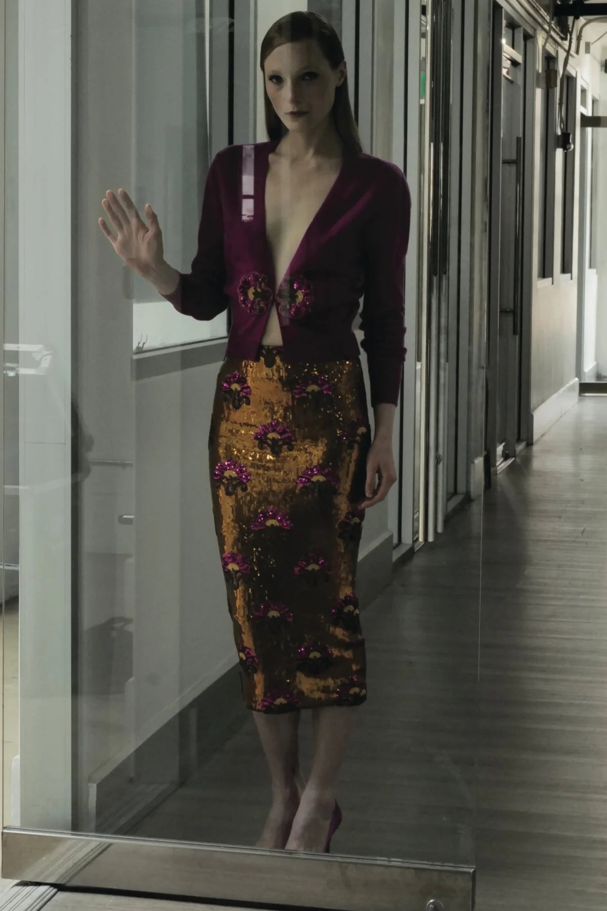 A woman with sleek, straight hair and a serious expression stands indoors behind a glass door, wearing the Le Superbe Liza Skirt—a figure-flattering sequin midi paired with a deep burgundy top—raising one hand to the glass.