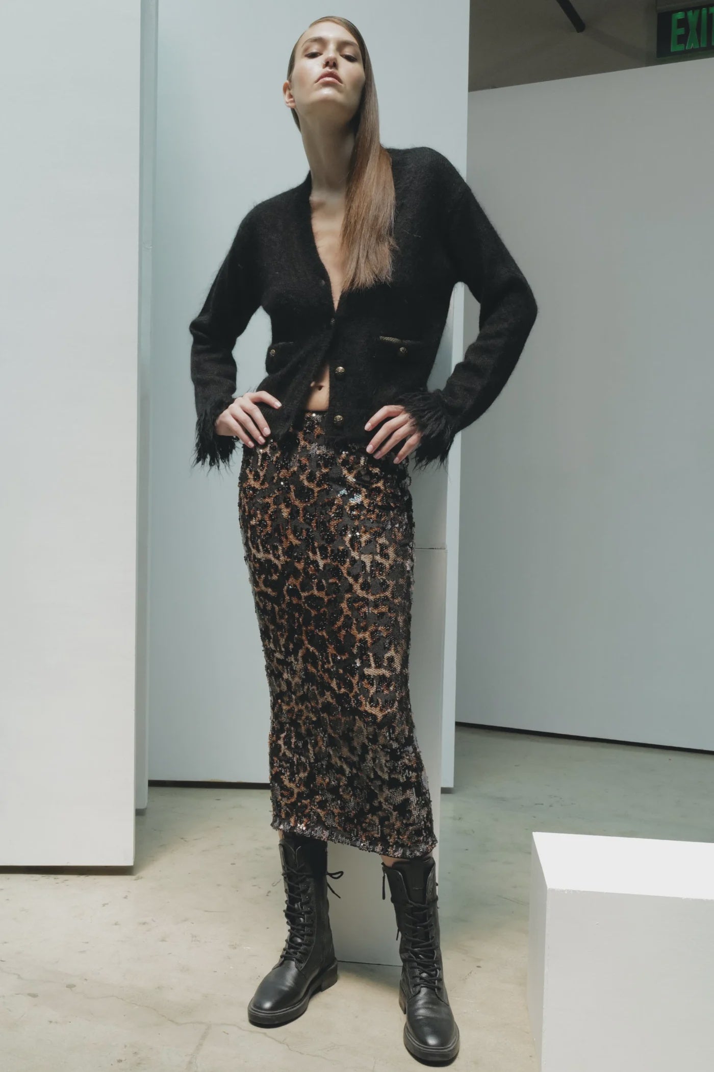 Indoors, a woman poses confidently in the Le Superbe Fresange Cardigan—a black, fuzzy textured knit button-up with feathered cuffs—paired with a leopard print sequin midi skirt and black lace-up combat boots. Her long hair is styled straight.