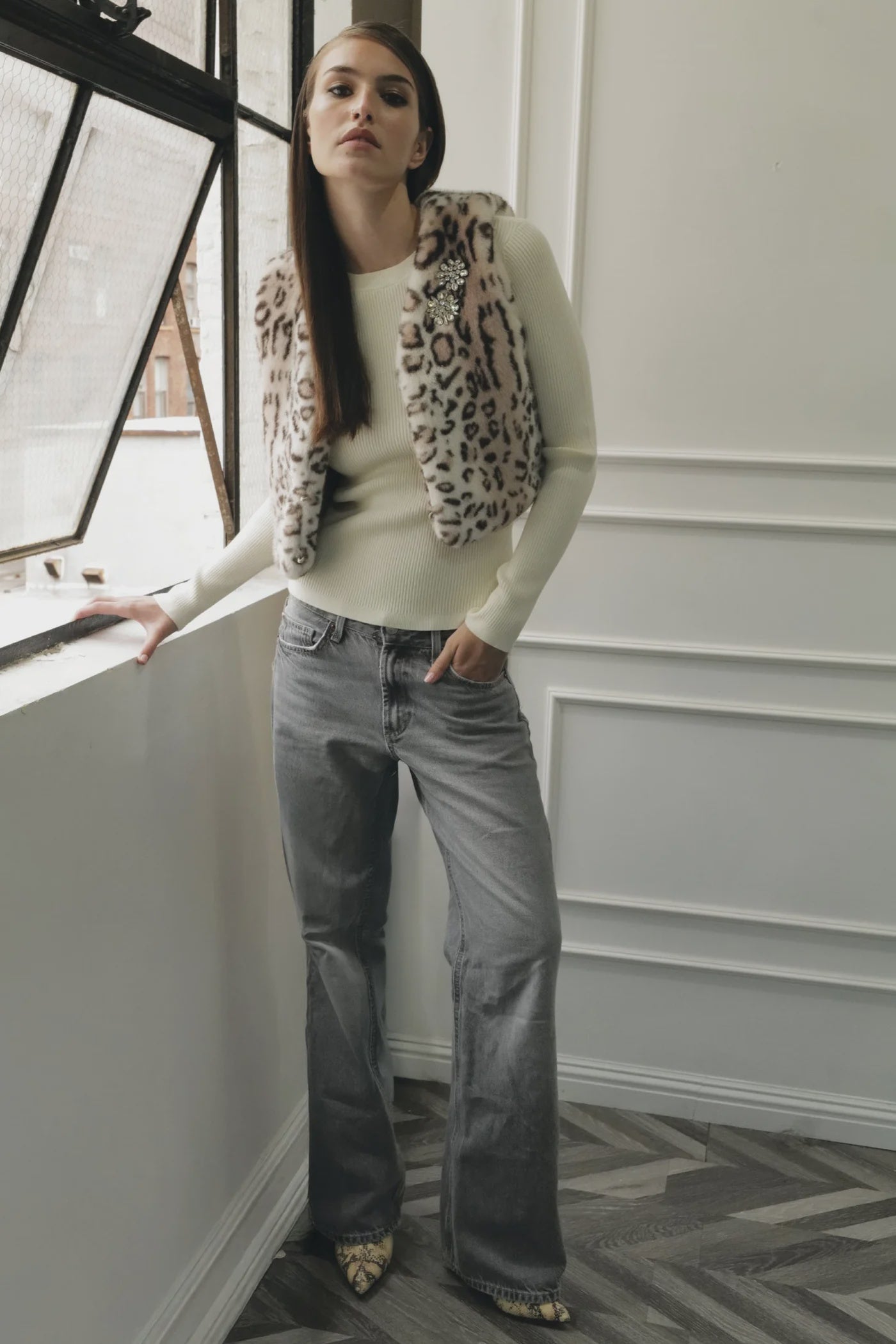 A woman with long straight hair stands indoors by a window, wearing Le Superbe’s Treasure Vest over a cream sweater, paired with gray flared jeans and snakeskin boots, one hand resting on the window ledge.