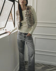 A woman with long straight hair stands indoors by a window, wearing Le Superbe’s Treasure Vest over a cream sweater, paired with gray flared jeans and snakeskin boots, one hand resting on the window ledge.