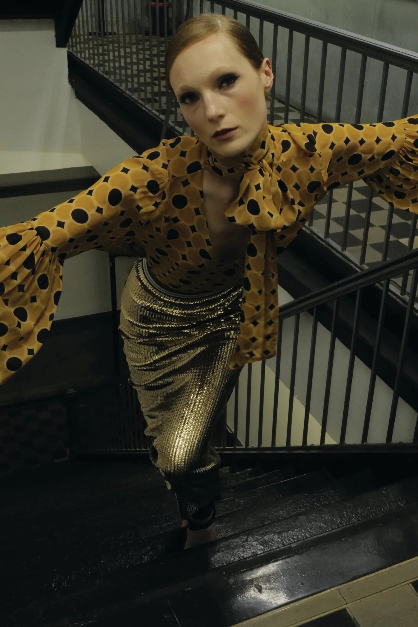Wearing the Le Superbe Serotonin Blouse, a person stands on a staircase with arms outstretched, posing dramatically and looking up toward the camera.