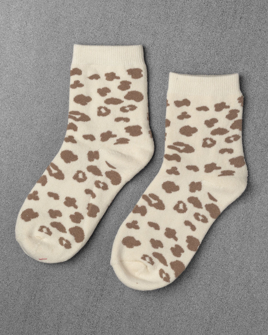 A pair of Unified Leopard Socks in cream with brown animal prints, laid flat on a textured gray surface.