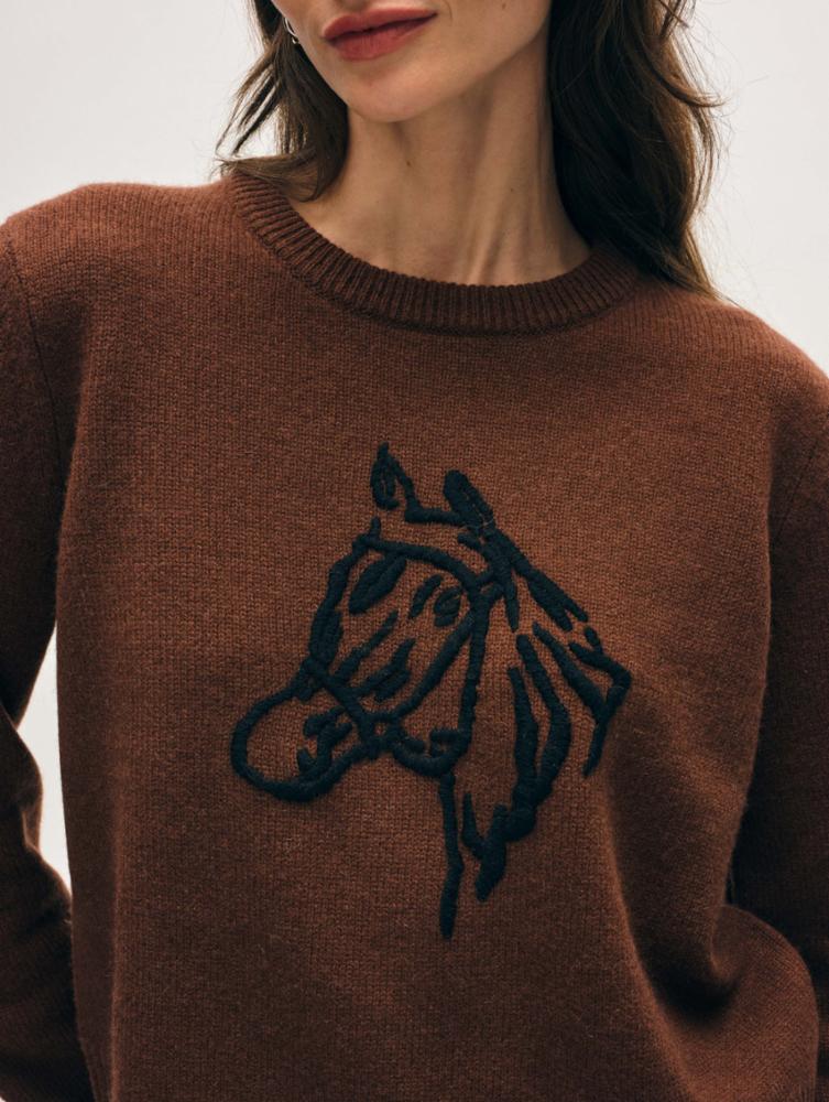 A woman models the White + Warren Horse Embroidered Crew, a brown Western-style sweater with a hand-embroidered horse's head in black. Only her lower face and upper torso are shown.