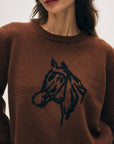 A woman models the White + Warren Horse Embroidered Crew, a brown Western-style sweater with a hand-embroidered horse's head in black. Only her lower face and upper torso are shown.