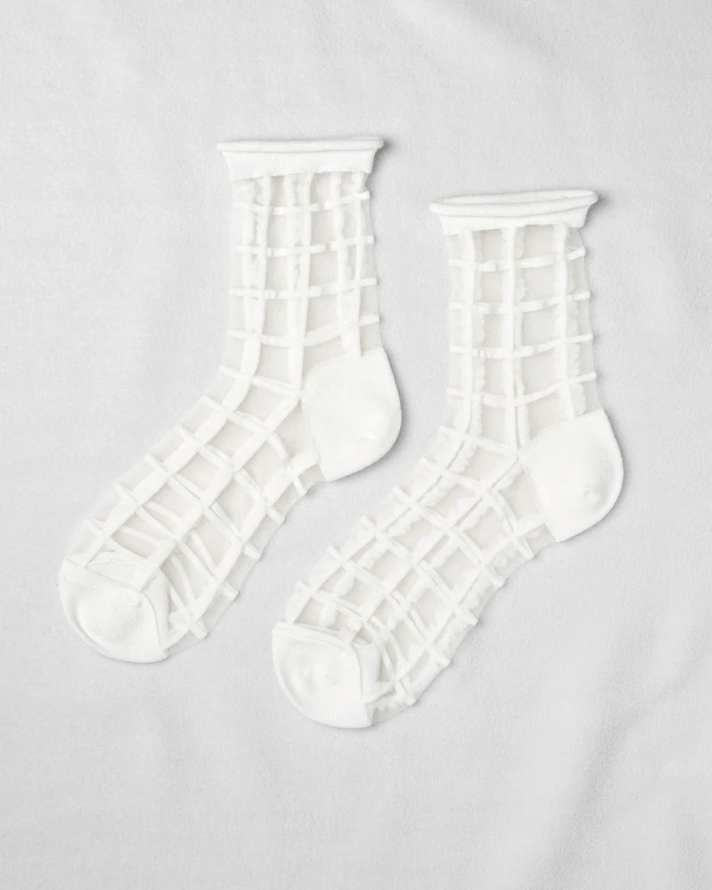 Unified’s Sheer French Window socks feature a white grid pattern on see-through fabric with solid white toes, heels, and cuffs for an airy look. Shown laid flat on a white surface.