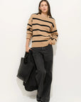 A woman stands against a white wall, holding a black textured tote bag. She wears Alex Mill’s Striped Nottinghill Sweater—an oversized, tan crewneck with black stripes—and loose black jeans.