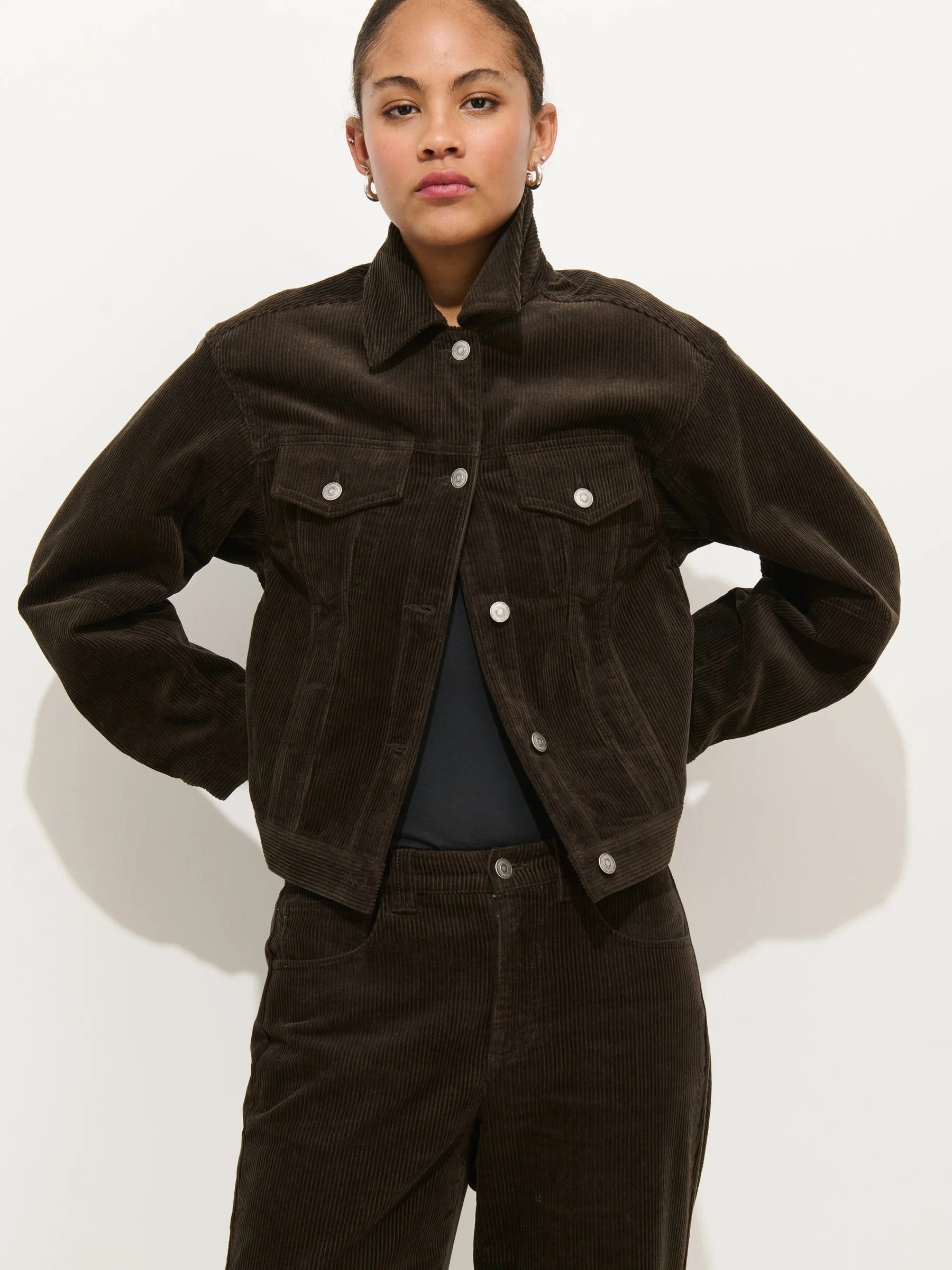 A person stands against a plain background wearing the Alex Mill Camden Rider Jacket in dark brown corduroy, paired with matching pants, hands on hips and a neutral expression.