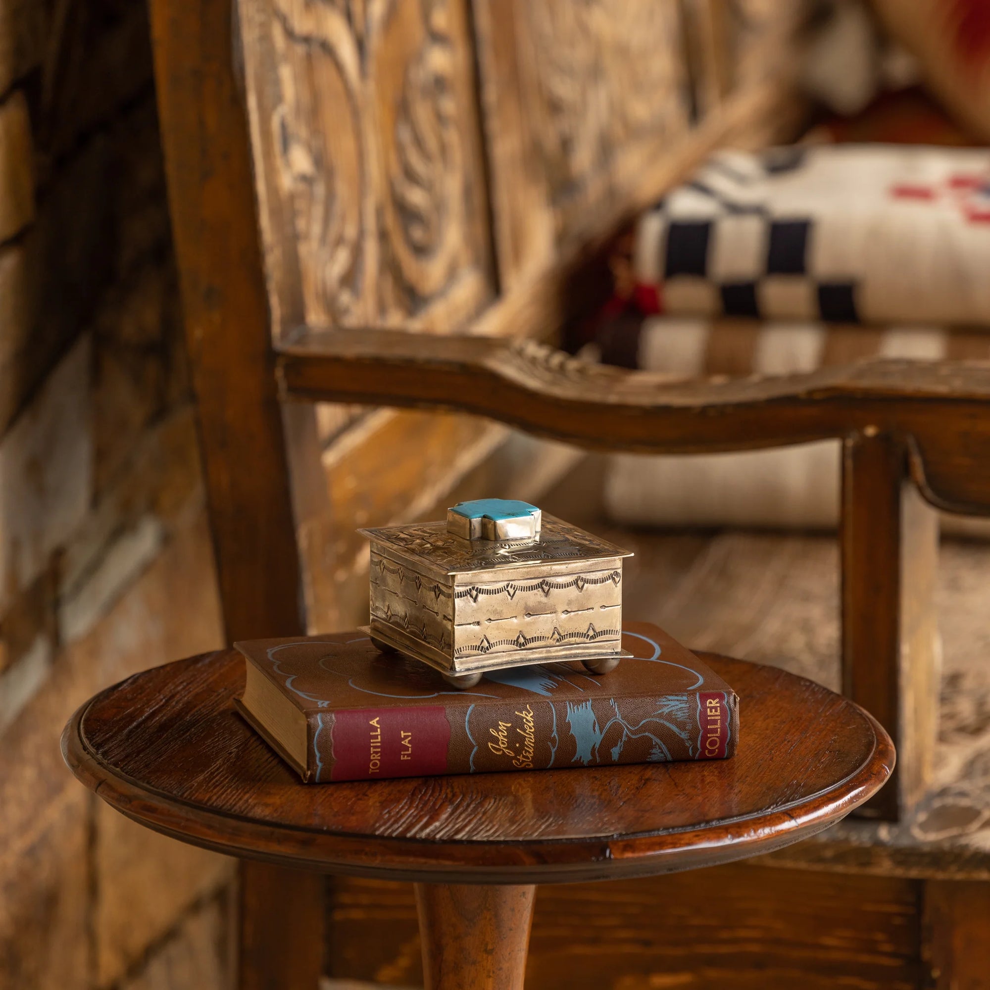 A Small Stamped Box w/ Turquoise Cross by J Alexander Rustic Silver and a hardcover book sit on a round wooden side table beside a carved bench with patterned cushions, bringing Southwestern charm to the cozy corner.