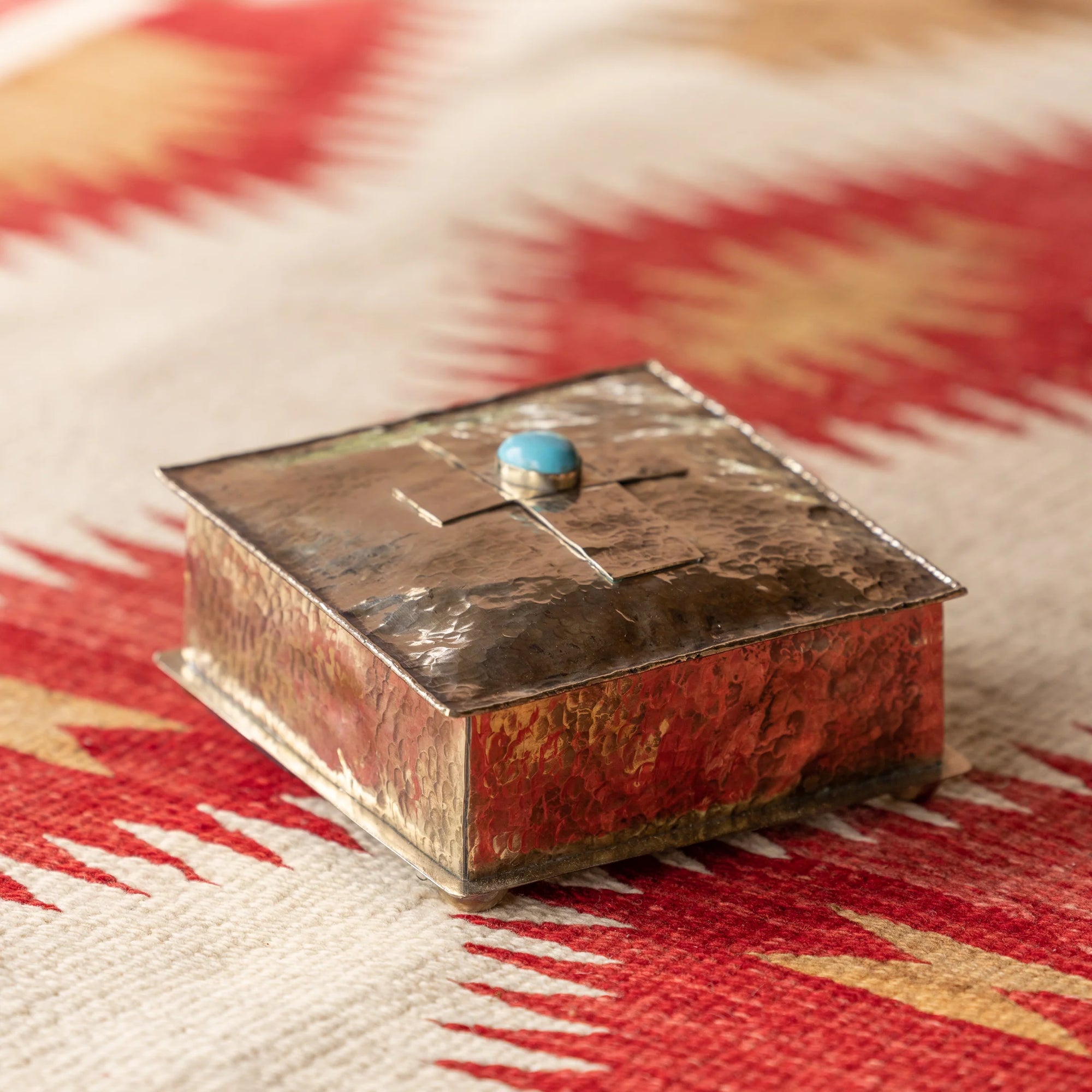 The SQ Silver Box w/ Dimples Cross by J Alexander Rustic Silver sits on a beige and red Southwestern-style geometric textile, featuring a hammered silver finish and a turquoise stone accent on its lid.