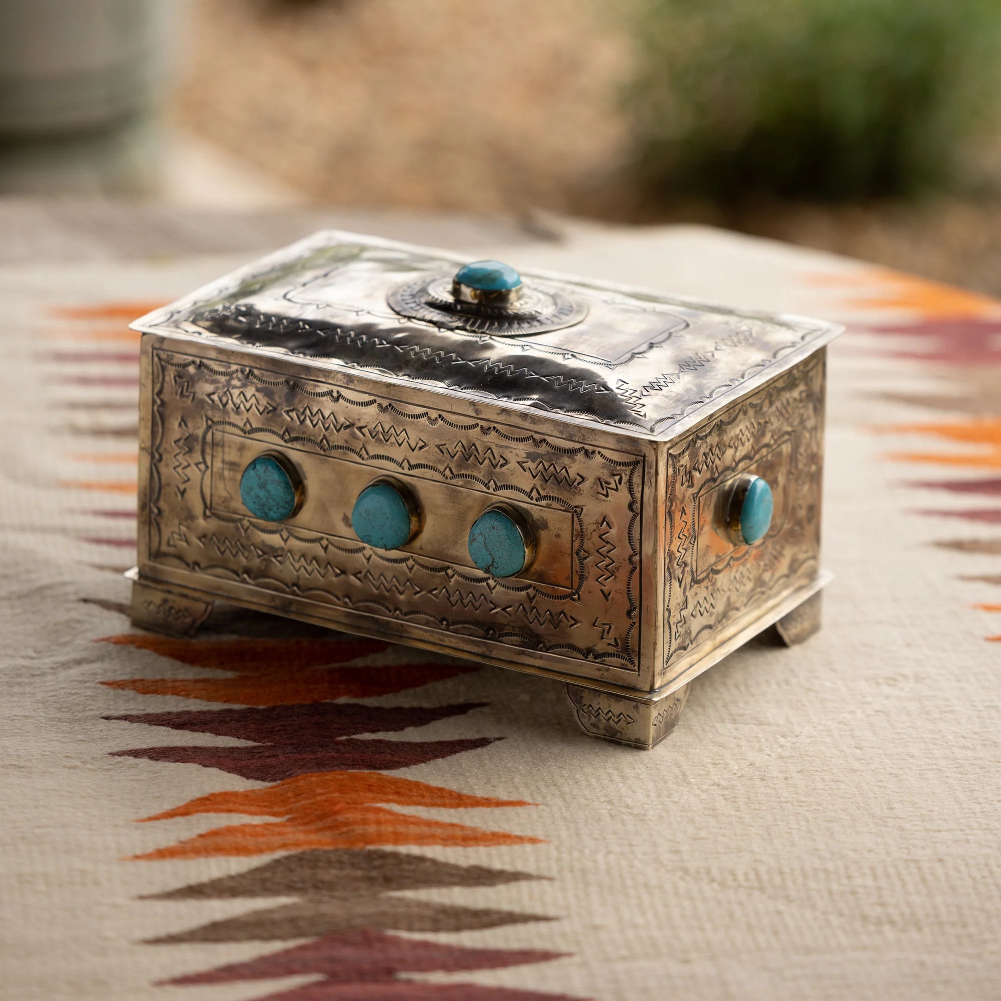 The J Alexander Rustic Silver 9 Stone Turq Box features engraved patterns and turquoise stones, displayed on a geometric woven fabric in brown, orange, and cream—perfect for adding a Southwestern touch to your space.