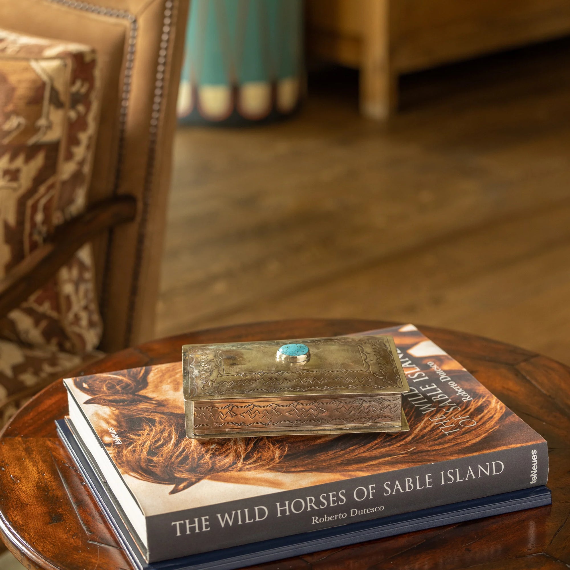 The Stamped Rec Box Turq by J Alexander Rustic Silver sits on "The Wild Horses of Sable Island" by Roberto Dutesco, bringing Southwestern style to a round wooden table next to an upholstered chair.