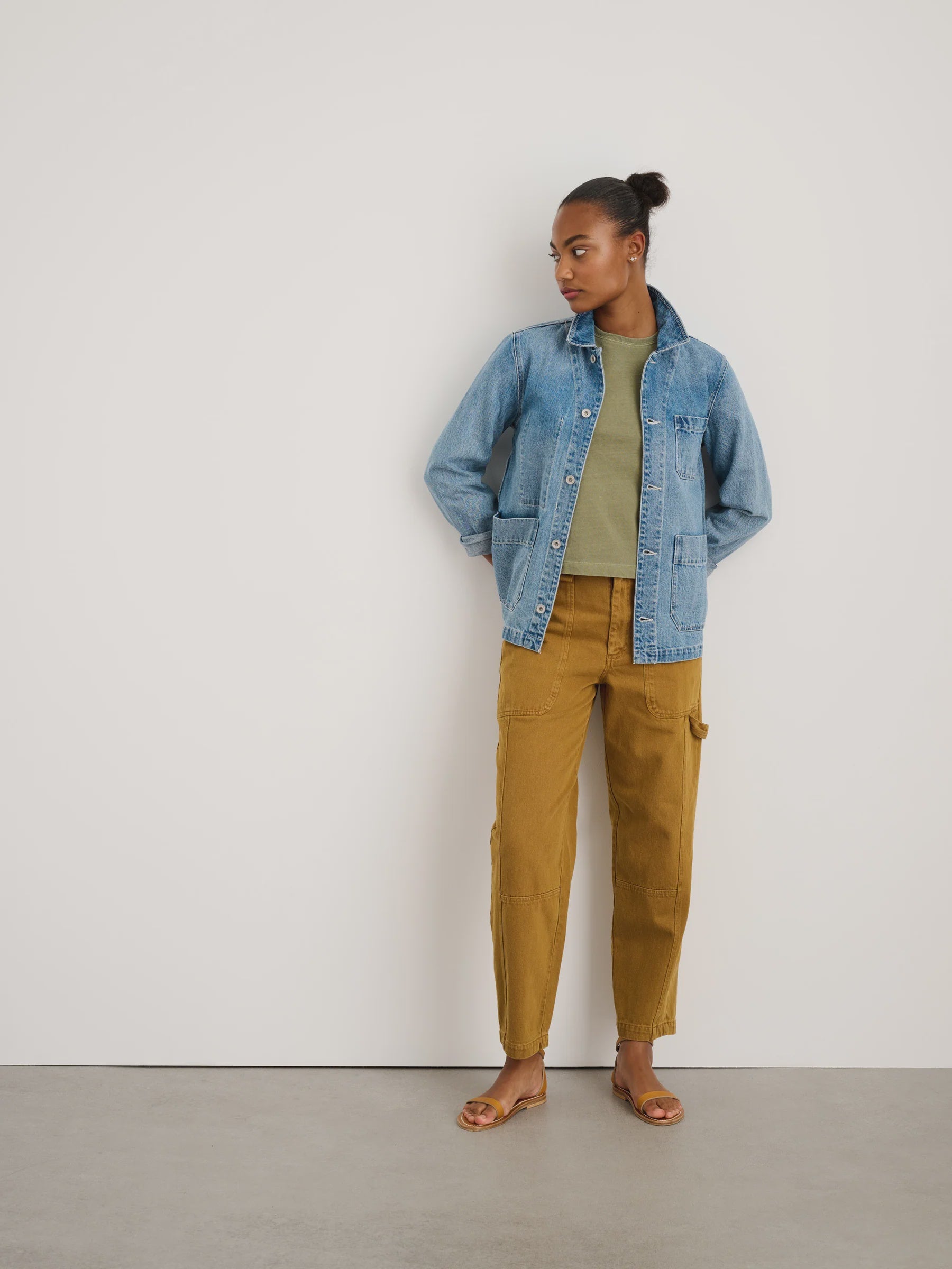 A woman stands against a white wall, wearing the Alex Mill Britt Work Jacket over an olive top, mustard pants, and tan sandals. She faces left with her hands behind her back.