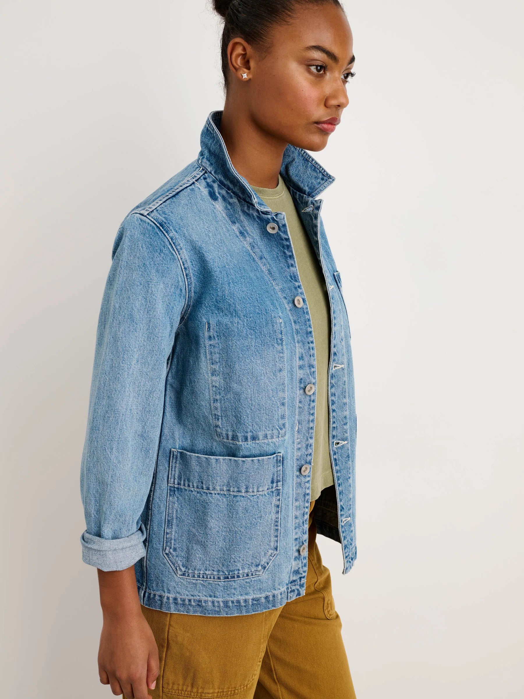 A person wears the Alex Mill Britt Work Jacket in light blue denim over a light green shirt and mustard pants, standing against a plain white background and looking to the side.