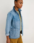 A person wears the Alex Mill Britt Work Jacket in light blue denim over a light green shirt and mustard pants, standing against a plain white background and looking to the side.