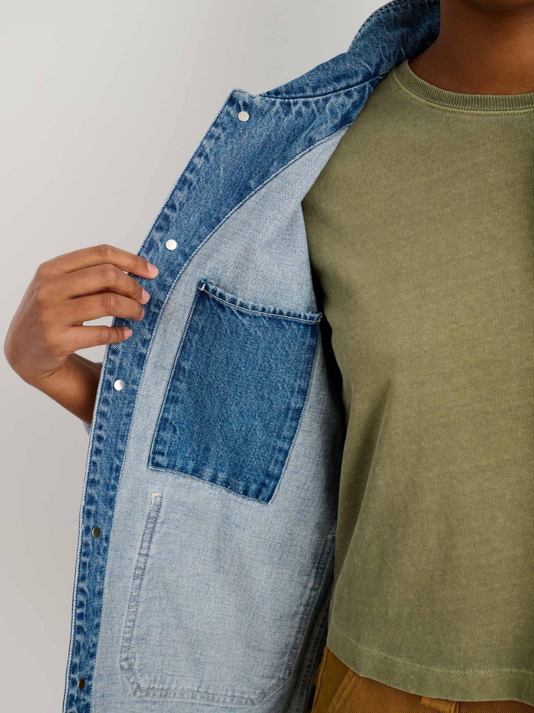 A person wearing an olive green t-shirt and brown pants opens an Alex Mill Britt Work Jacket in blue recycled denim, revealing the inside left pocket.