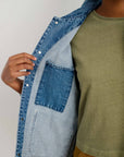 A person wearing an olive green t-shirt and brown pants opens an Alex Mill Britt Work Jacket in blue recycled denim, revealing the inside left pocket.