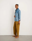 A woman stands sideways against a white wall, wearing the Alex Mill Britt Work Jacket in light blue recycled denim, mustard yellow wide-leg pants with pockets, and brown flat sandals. Her hair is pulled back as she looks at the camera.