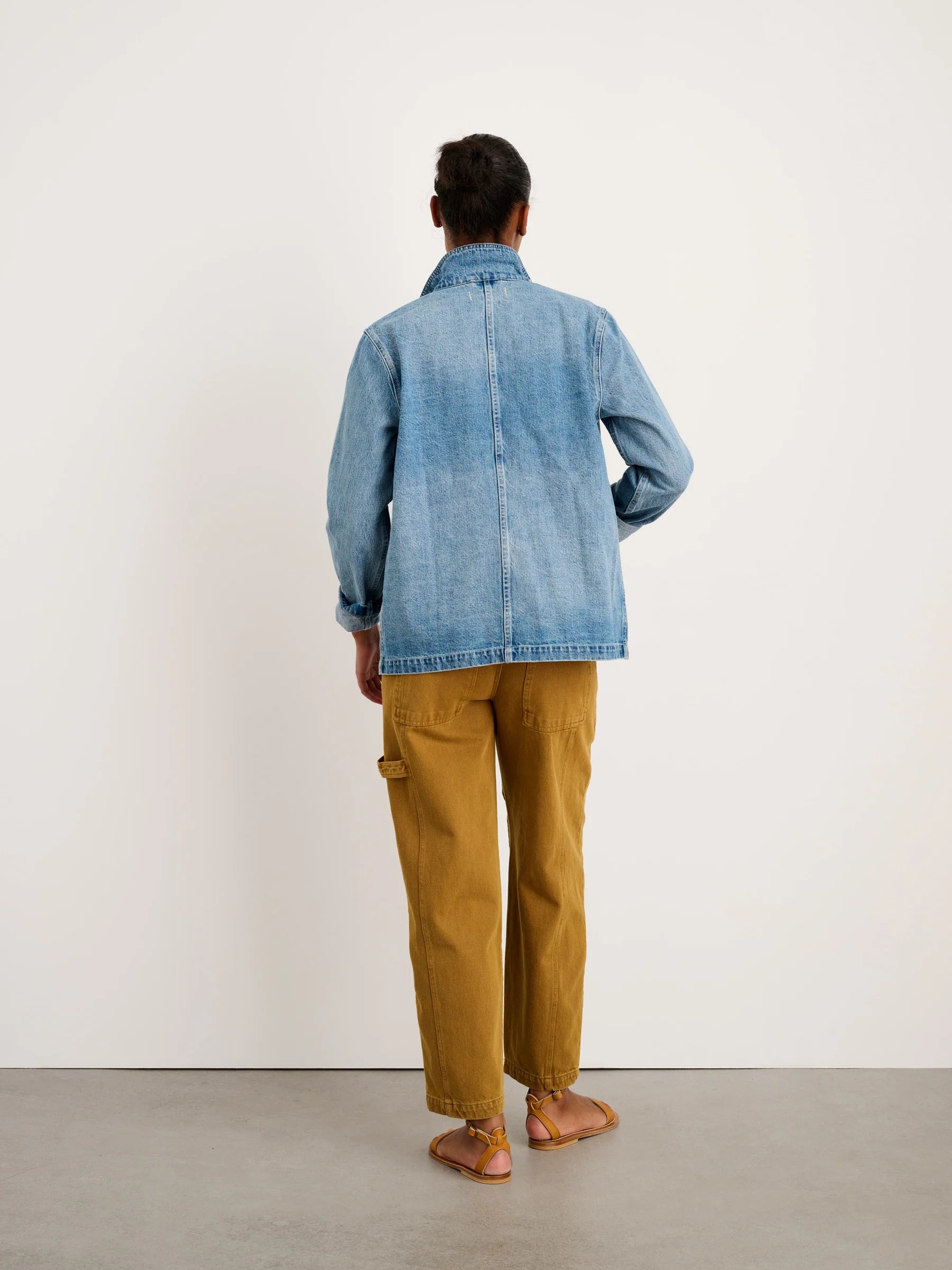 A person stands with their back to the camera wearing the Britt Work Jacket by Alex Mill, tan cargo pants, and brown sandals against a plain white wall.