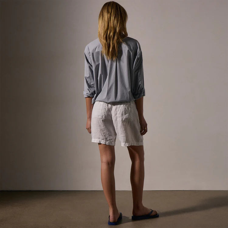 A person with long hair faces away from the camera, wearing a loose gray long-sleeve shirt, James Perse Lightweight Linen Short in white with an elastic waistband, and sandals against a plain background.