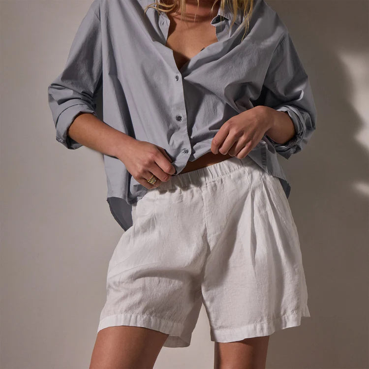 A person stands against a neutral background wearing James Perse Lightweight Linen Shorts with an elasticized waistband and a loose, light blue button-up shirt with rolled sleeves; their face is not visible.