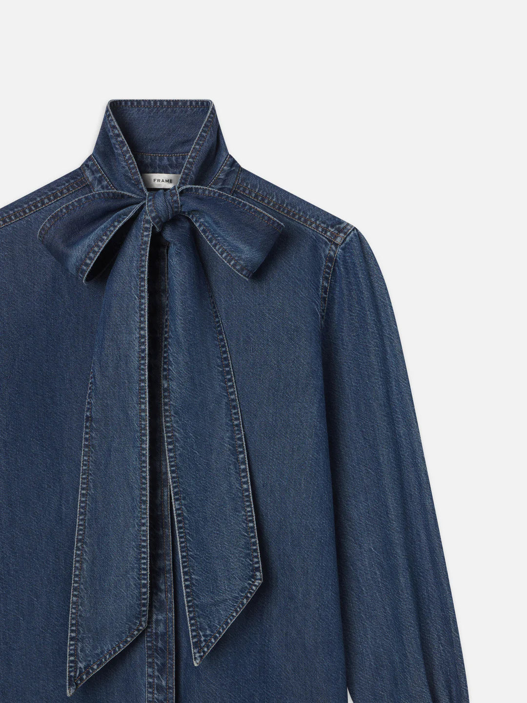 The FRAME Secretary Blouse is a stylish denim shirt with long sleeves and a bold bow at the collar, providing a contemporary update to the traditional design.