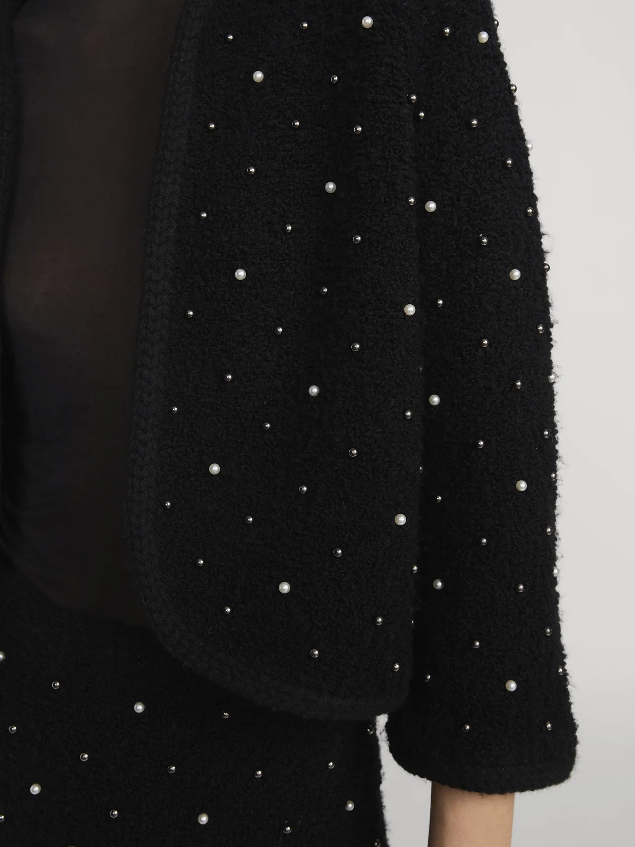 A close-up of a person wearing FRAME's The Embellished Jacket, a black textured knit adorned with small pearls and beads over a sheer top. The light background highlights every detail of this striking piece.