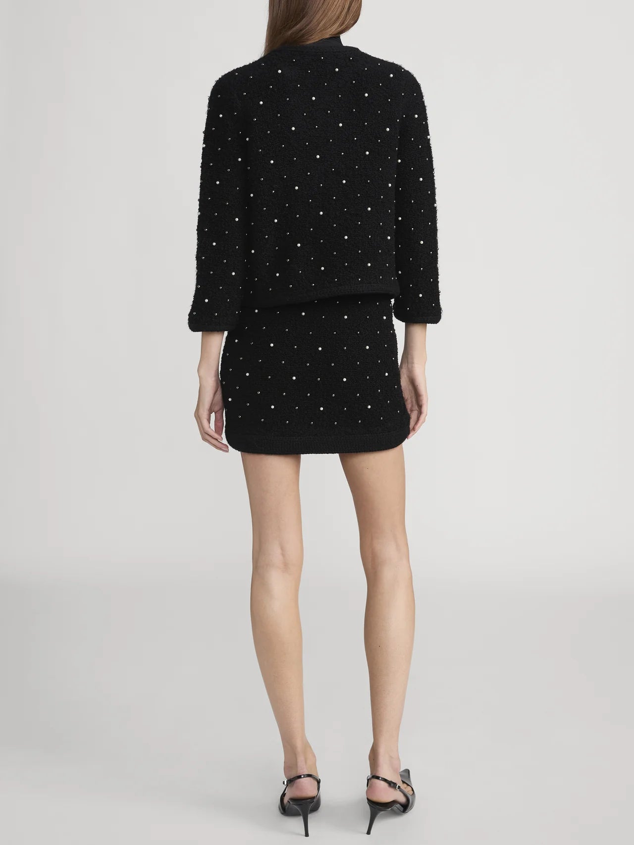 A woman faces away, wearing FRAME's The Embellished Jacket—a black textured knit with small white pearls—paired with a matching short skirt and black heeled sandals. The background is plain light gray.